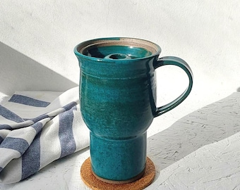 May include: A teal ceramic mug with a handle and a lid, sitting on a round cork coaster. The mug has a cylindrical base and a wider top, with a dark interior. A blue and white striped cloth is partially visible in the background.