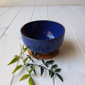 May include: A deep blue ceramic bowl with a glossy finish, resting on a cork coaster. The bowl's interior reflects light, and a sprig of green leaves with yellow tips adds a natural touch. The bowl is a decorative item.