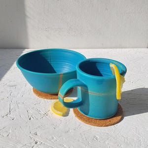 以下が含まれることがあります： ターコイズブルーのセラミックボウルとマグカップのセット。レモンスライスが添えられています。マグカップには取っ手があり、コルクコースターの上に置かれています。ボウルはマグカップの隣にあります。