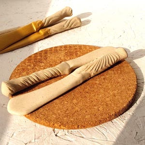 Può includere: Due coltelli da burro in ceramica beige con manici strutturati poggiano su un sottopentola rotondo in sughero. Uno ha un design a spirale, l'altro un motivo a raggiera. È visibile anche un paio di coltelli da burro color oro.
