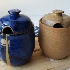 May include: Two ceramic salt cellars with lids. One is blue with a black stripe and a tan stripe. The other is tan. Both have a hole in the lid for pouring.