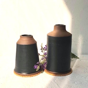 Peut inclure: Deux vases en céramique avec un corps bleu foncé et un bord marron. Les vases sont cylindriques et reposent sur une base ronde en liège. Des fleurs violettes sont placées entre les vases. Les vases sont faits à la main et ont une esthétique rustique.