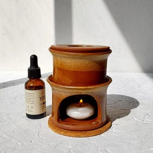 May include: A ceramic essential oil diffuser in shades of brown with a lit candle inside. A small brown bottle with a dropper is to the left. The diffuser sits on a cork base.