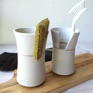 Può includere: Due supporti cilindrici in ceramica bianca con finitura opaca e una linea orizzontale incavata al centro. Un supporto contiene una spugna metallica dorata, l'altro presenta una fessura verticale che sostiene una spazzola per piatti bianca.