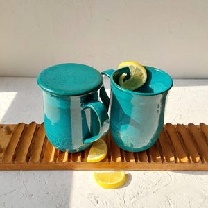 Puede incluir: Dos tazas de cerámica azul turquesa con una tapa en una de las tazas. Las tazas están sobre una bandeja de madera. Una rodaja de limón está en una de las tazas.