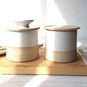 以下が含まれることがあります： 蓋付きのセラミックバターディッシュ2つ。 各皿の上半分は白く、下半分はまだらのベージュです。 1つの蓋にはハンドルが付いています。 皿は木製のまな板の上にあります。