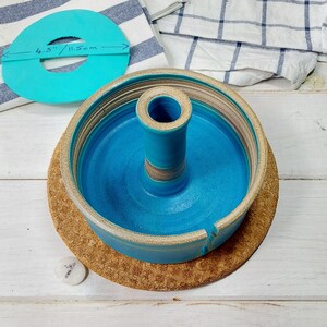 May include: A turquoise ceramic cake stand with a central cylinder and a 4.5 inch diameter. The stand is on a cork trivet.