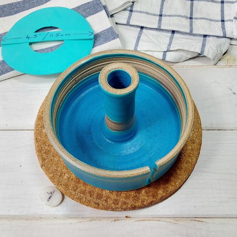 May include: A turquoise ceramic cake stand with a central cylinder and a 4.5 inch diameter. The stand is on a cork trivet.