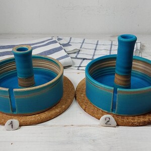 May include: Two ceramic yarn bowls with a center post for holding yarn. The bowls are teal blue with brown stripes and are sitting on cork coasters. The coasters are labeled with the numbers 1 and 2.