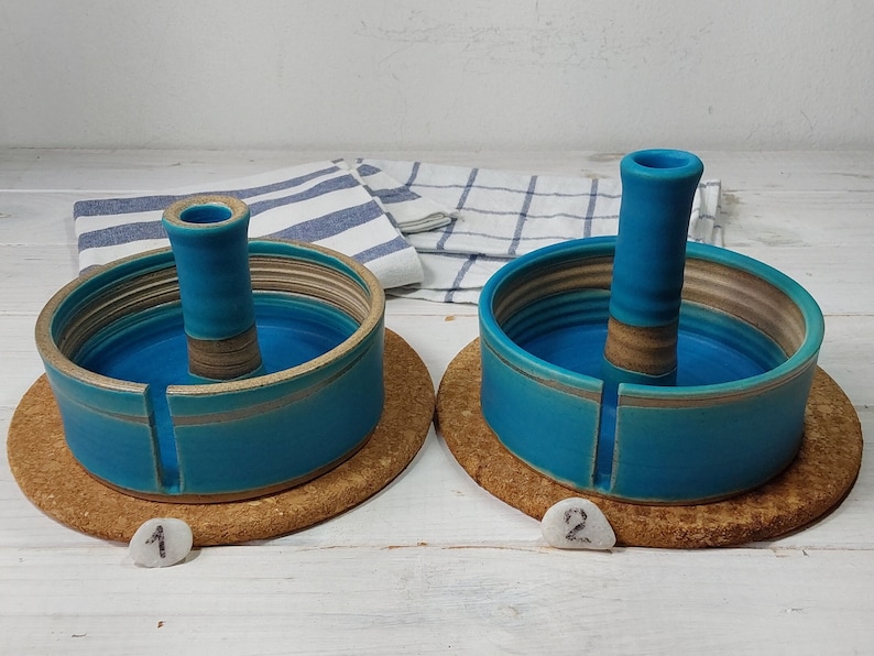 May include: Two ceramic yarn bowls with a center post for holding yarn. The bowls are teal blue with brown stripes and are sitting on cork coasters. The coasters are labeled with the numbers 1 and 2.