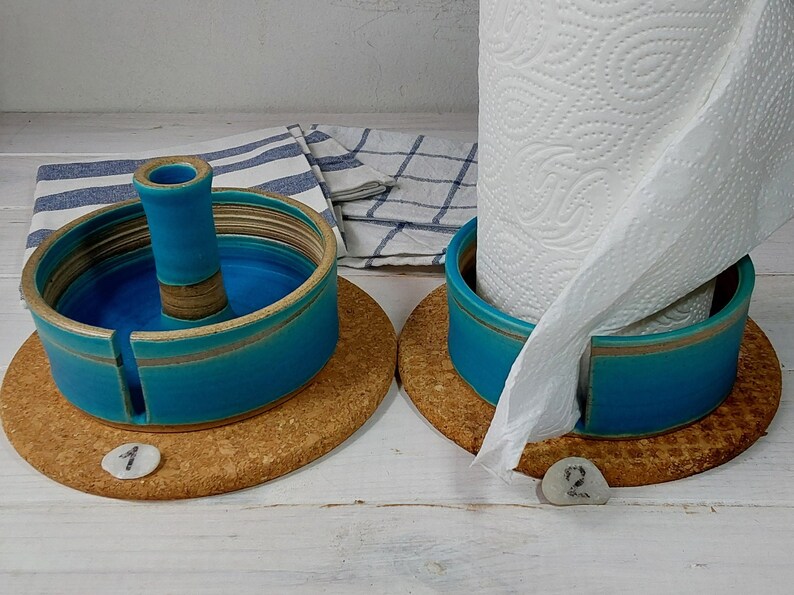 May include: Two ceramic paper towel holders with cork bases. The holders are teal blue with a light brown stripe. One holder has a paper towel roll inside. Both holders have a number on the base, one is number 1 and the other is number 2.