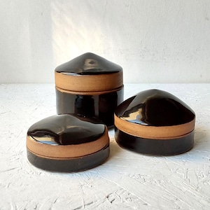 May include: Three cylindrical ceramic containers with lids, featuring a glossy dark brown glaze on the top and bottom sections, contrasted by an exposed matte tan clay band around the centre of each piece, arranged in a cluster on a white surface.