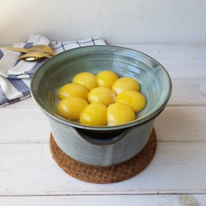 May include: A ceramic bowl set with a light blue glaze, containing bright yellow egg yolks. The bowls sit on a cork coaster. A blue and white striped cloth and gold serving utensils are in the background. A kitchenware item.