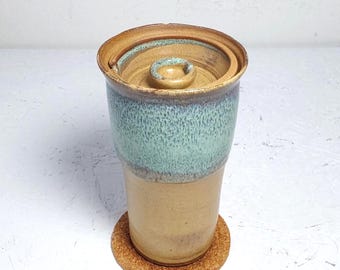 May include: A cylindrical ceramic container with a lid, featuring a smooth tan base and a textured, speckled light green glaze on the upper portion. The lid has a small central knob, and the entire piece rests on a circular cork coaster.