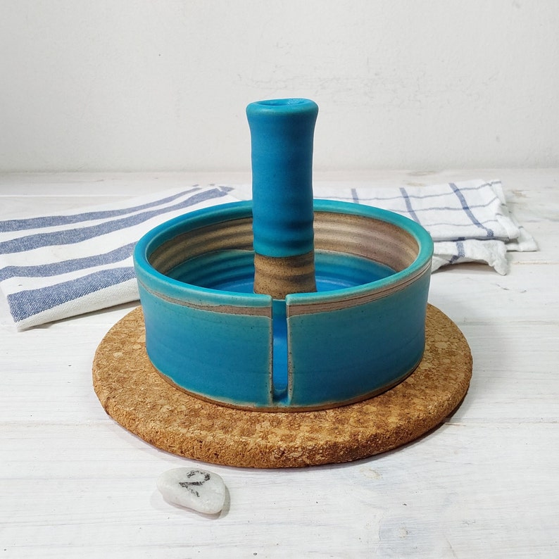 May include: A teal blue ceramic yarn bowl with a center post and a cork base. The bowl has a light brown stripe around the top and bottom.