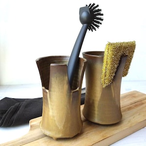 May include: Two ceramic kitchen utensil holders in shades of brown and gold. One holds a black handled brush with black bristles, the other a gold scrubby. The holders sit on a wooden cutting board.