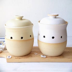 Puede incluir: Dos recipientes de cerámica para ajo con tapas. Uno es de color crema, el otro blanco. Ambos tienen una banda de color canela en la base y agujeros para ventilación. Están sobre una tabla de cortar de madera.