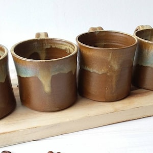 May include: Four brown ceramic mugs with a light blue and yellow glaze. The mugs are arranged in a row on a wooden surface.