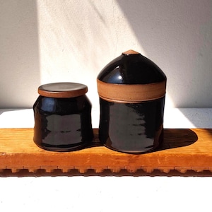 May include: Two ceramic canisters with glossy black glaze and exposed natural clay rims. The shorter canister has a flat lid, while the taller canister features a domed lid with a pointed top, both showcasing a smooth, reflective finish.