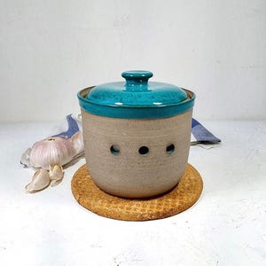 May include: A ceramic garlic keeper with a teal lid and rim, and a tan body with three ventilation holes. The keeper sits on a round, textured cork coaster. A bulb of garlic is visible nearby.