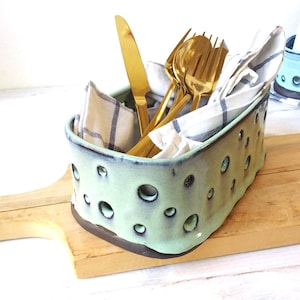 Könnte beinhalten: Ein hellgrüner Keramik-Utensilienhalter mit unregelmäßigen Löchern, der goldfarbenes Besteck und ein weiß-blau gestreiftes Tuch enthält. Der Halter steht auf einem Holzschneidebrett und schafft eine rustikale Küchenästhetik.