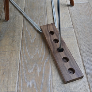 Può includere: Un supporto in legno per tappi di bottiglia di vino con quattro aperture circolari. Un tappo di bottiglia di vino in metallo è inserito in uno dei fori. Il supporto è collegato a una corda e poggia su un pavimento in legno chiaro.