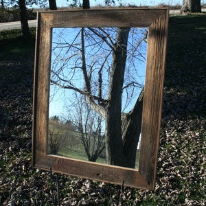 May include: A rustic wooden mirror with a weathered brown frame. The mirror reflects a tree with branches reaching towards the sky.