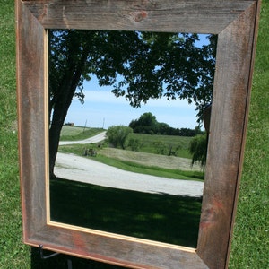 May include: A rustic wooden mirror with a weathered brown frame and a clear glass reflection of a country road and trees. The mirror is standing on a black metal easel.