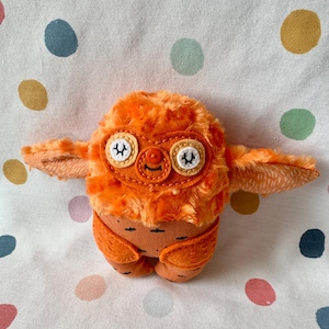 May include: A handmade orange plush creature with a fluffy head, stitched eyes, and small wings. The plush toy has a carrot-shaped body and is set against a polka dot background.