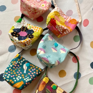 May include: Five small fabric pouches with straps, each featuring a unique pattern including floral, geometric, vegetable, and acorn prints. Each pouch is secured with a single plastic snap button in colours like pink, green, and blue.