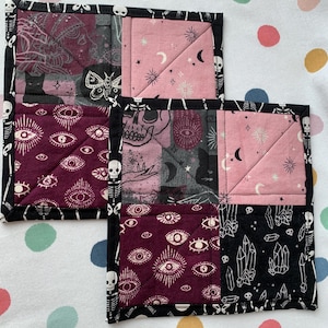 May include: Two square quilted coasters with a black skeleton border. Each coaster features a patchwork design with various patterns, including eyes, moons, skulls, and crystals. The color palette includes burgundy, pink, black, and white.