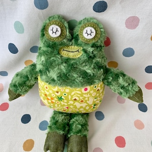 May include: A plush green frog toy with a textured, furry body. The frog has large, round eyes with white accents and stitched eyelashes. Its belly is a light green fabric with a floral pattern. The frog's feet and arms are a darker green corduroy.