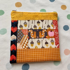May include: A small, square quilted pouch with a zipper closure. The pouch features a patchwork design with various fabrics, including a yellow sunburst pattern, squirrels, acorns, and strawberries. The color palette includes shades of yellow, orange, red, and brown.