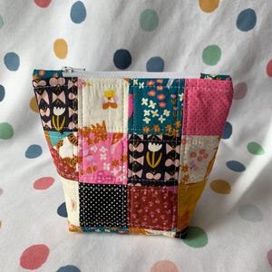 May include: A small, patchwork fabric pouch with a white zip. The pouch is made of various patterned squares in colours like pink, teal, yellow, and black. The patterns include floral designs, polka dots, and geometric shapes. The pouch is on a white surface with colourful polka dots.
