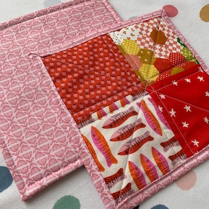 May include: Two handmade quilted square hot pads. One pad features a pink floral pattern, while the other has a patchwork design with red, white, and pink fabrics. The patchwork includes fish, stars, and geometric shapes.