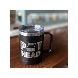 May include: Black insulated coffee mug with a silver rim and handle. The mug features the words "POT HEAD" in a bold, white, sans-serif font, with a coffee pot graphic replacing the "O". The mug has a clear, plastic lid.