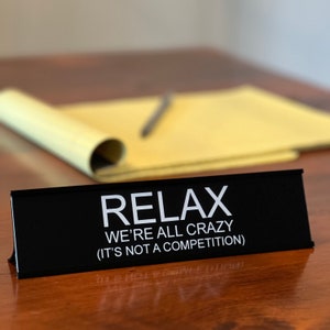 May include: Black desk nameplate with white text that reads "Relax We're All Crazy (It's Not A Competition)"