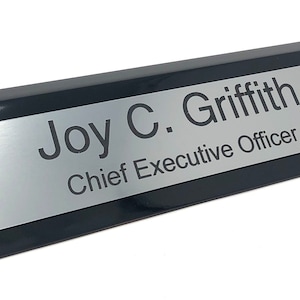 May include: Black desk nameplate with a silver insert. The nameplate reads "Joy C. Griffith, Chief Executive Officer."