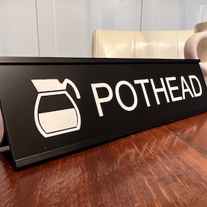 May include: Black desk sign with white text that reads "POTHEAD" and a white silhouette of a coffee pot.