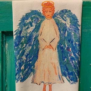 May include: A white tea towel with a blue and green angel painting. The angel has a halo and is holding its hands together. The words "Peace" are written in blue below the angel.