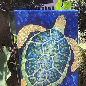 May include: A colorful garden flag featuring a green, blue, and yellow sea turtle swimming in a blue ocean. The turtle's shell is detailed with a pattern of geometric shapes. The flag is printed with the artist's signature, "Gerry", in the bottom right corner.