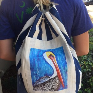 May include: A white and blue striped canvas tote bag with a pelican illustration. The pelican is brown and gray with a long orange beak. The bag has a white background with a blue stripe pattern.