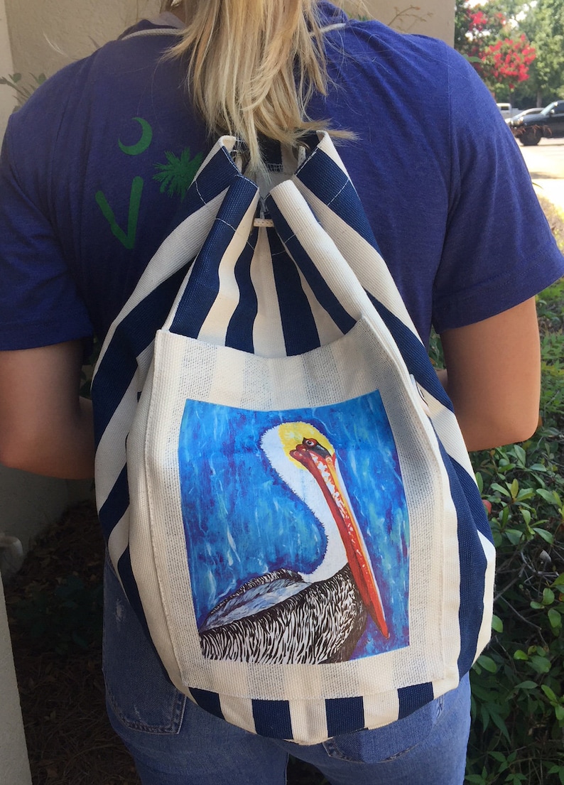 May include: A white and blue striped canvas tote bag with a pelican illustration. The pelican is brown and gray with a long orange beak. The bag has a white background with a blue stripe pattern.