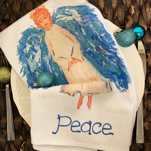 May include: White kitchen towel with a watercolor painting of an angel with blue wings and the word "Peace" printed in blue below the angel.