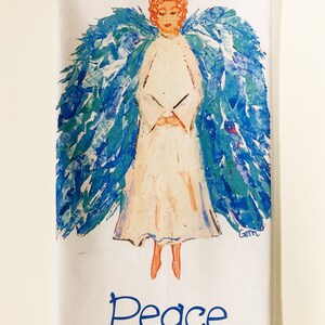 May include: White kitchen towel with a blue and green angel illustration and the word "Peace" in blue lettering. The angel has a halo and is painted in a whimsical style.