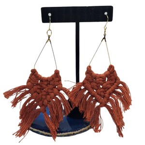 May include: A pair of terracotta-colored macrame earrings with fringe hanging from the bottom. The earrings are displayed on a black stand.