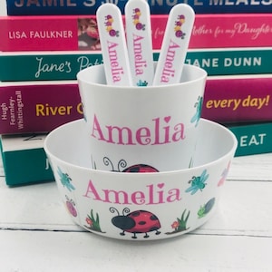 May include: A white plastic bowl and cup with a ladybug and dragonfly design. The bowl and cup are personalised with the name "Amelia" in pink lettering.