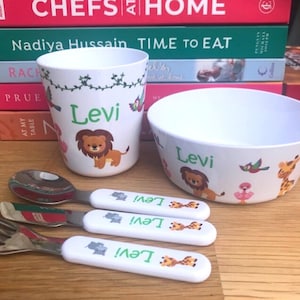 May include: A white children's dinner set including a cup, bowl, spoon, and fork. The set is decorated with animal illustrations, including a lion and giraffe, and the name "Levi" in green. Cookbooks are visible in the background.