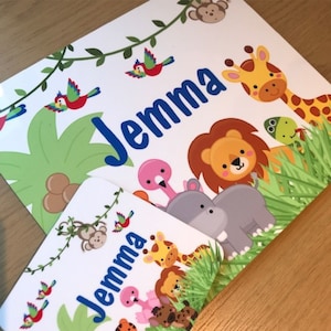 May include: Two white rectangular prints with a jungle theme. The prints feature the name "Jemma" in blue, along with illustrations of a giraffe, lion, hippo, and other jungle animals. Green leaves and vines are also present.