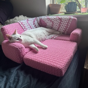May include: A white cat lounges on a small, pink crocheted sofa with a matching pink and white crocheted blanket. The sofa is placed on a bed with a dark blue comforter. The cat is the focal point of the image.